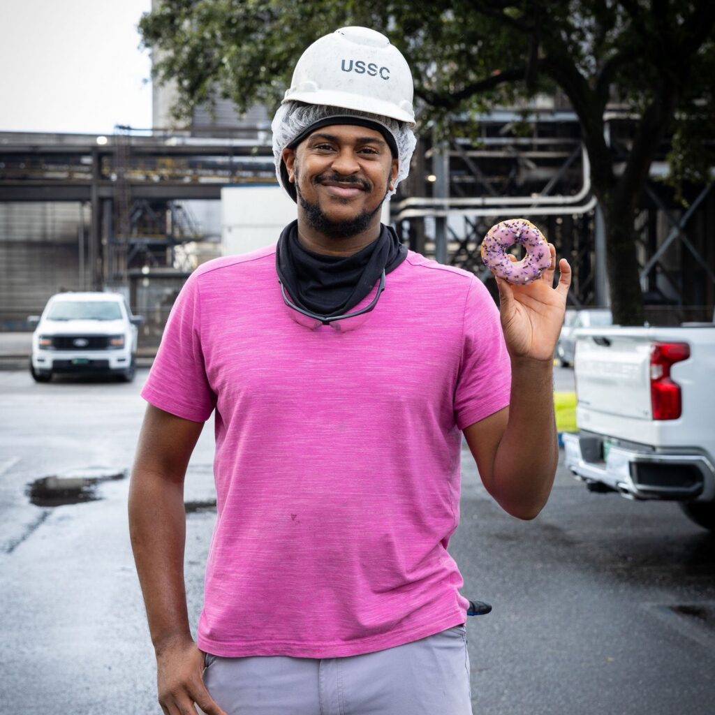 A U.S. Sugar employee wearing a pink shirt and hard hat holds up a pink-frosted doughnut in recognition of Breast Cancer Awareness Month.