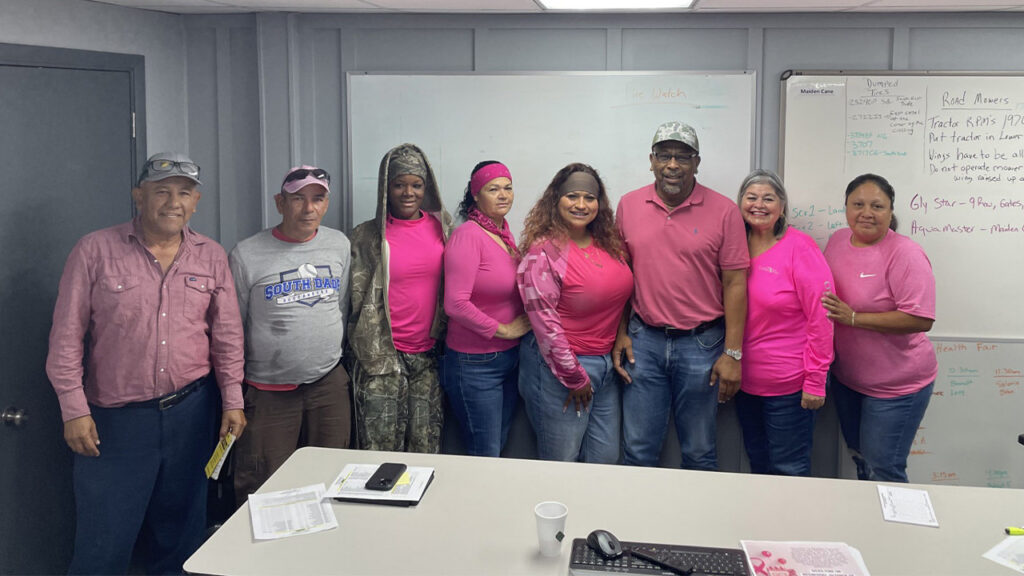 Eight U.S. Sugar employees, many dressed in pink, gather in a meeting room to show support for breast cancer awareness.
