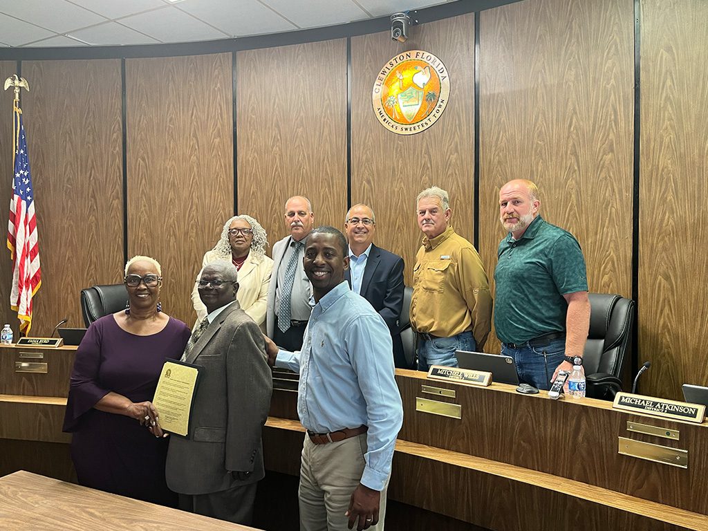A group of community leaders and officials stand in a wood-paneled county commission chamber in Clewiston, Florida, as Brannan Thomas smiles in the foreground while a certificate of recognition is presented, honoring his role in supporting the Harlem Tree Park Amphitheater project.