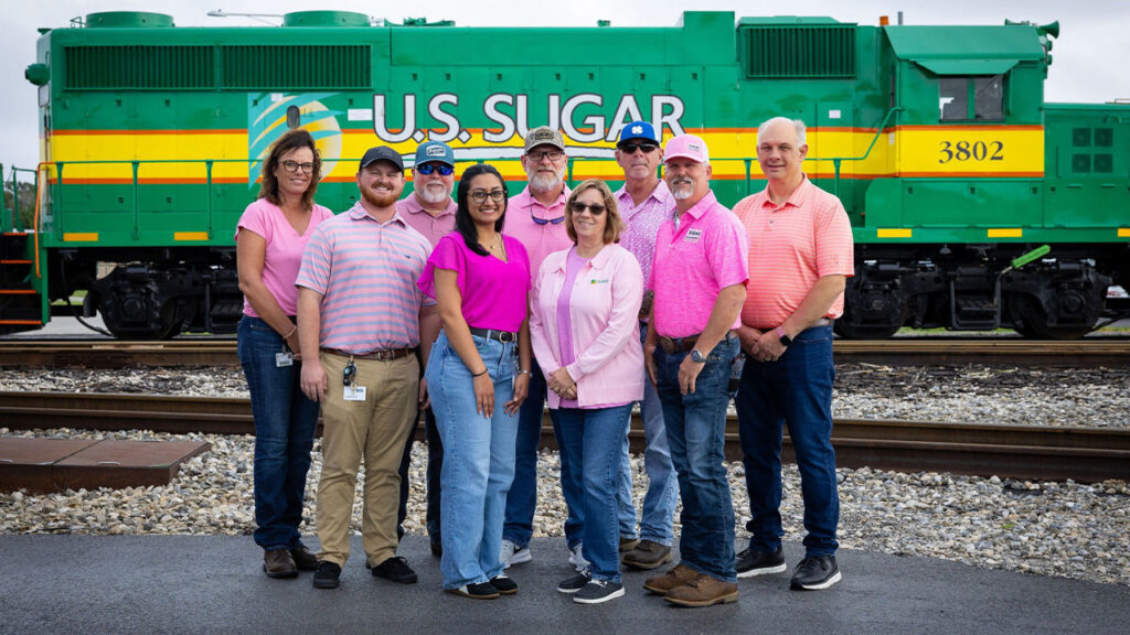 Nine U.S. Sugar employees wearing shades of pink pose in front of a green and yellow U.S. Sugar locomotive for Breast Cancer Awareness Month.