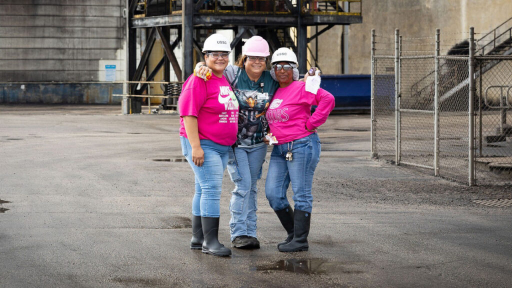 Three U.S. Sugar employees wearing pink clothing and hard hats pose together at an industrial facility in support of Breast Cancer Awareness Month.