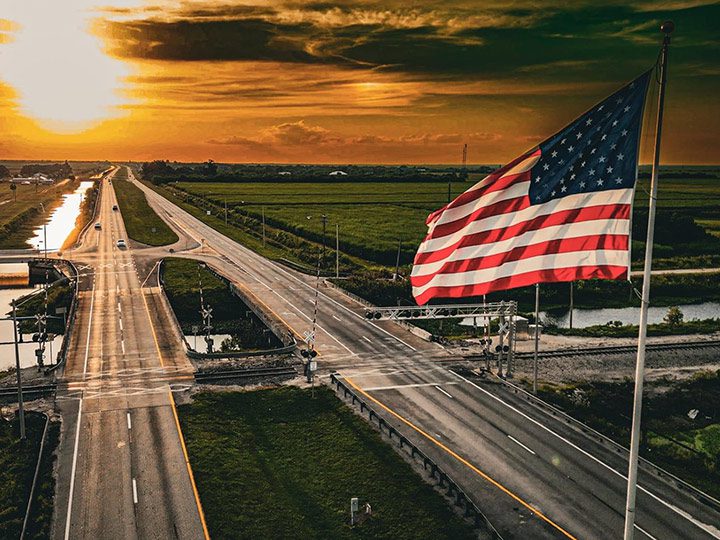 American flag waving beside a rural roadway and railroad crossing at sunset, representing U.S. Sugar’s role in American agriculture, rural communities and the nation’s America 250 celebration.