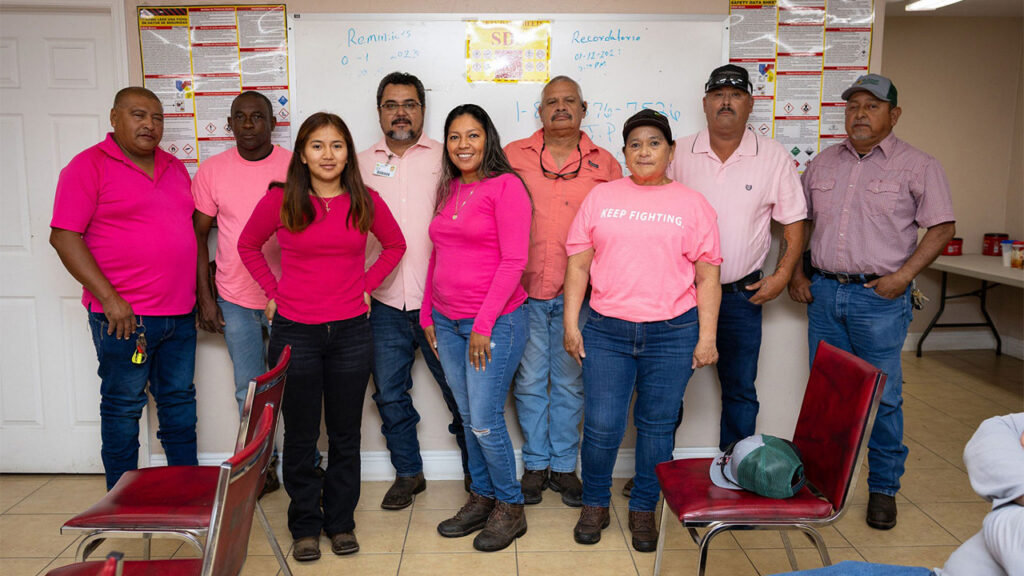Nine U.S. Sugar employees wearing pink shirts and other pink clothing pose together indoors in support of breast cancer awareness.